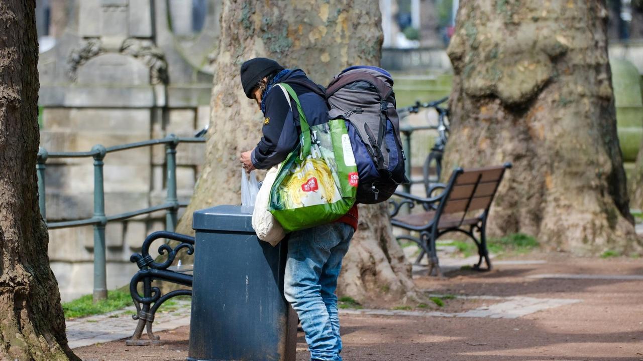 Ein Mann mit Turnschuhen und schlecht sitzenden Jeans durchsucht einen Abfalleimer an einer Parkbank. Er trägt mehrere große Plastiktaschen und einen Rucksack über der Schulter.