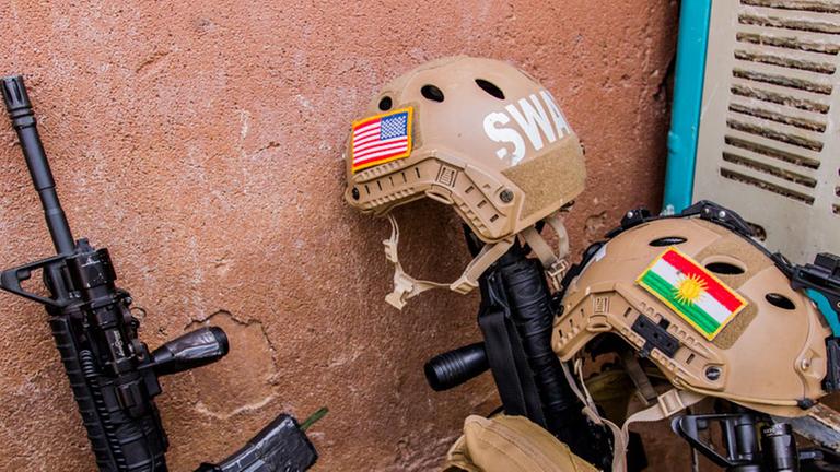 An einer Wahn lehnen Gewehre, daneben Helme mit den Flaggen Iraks und der USA.