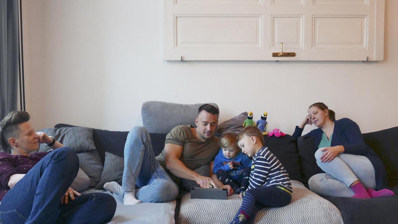 Eine Familie, bestehend aus zwei Müttern, einem Vater und zwei Kindern sitzen zusammen auf der Couch. Die Kinder schauen mit dem Vater auf ein Tablet.