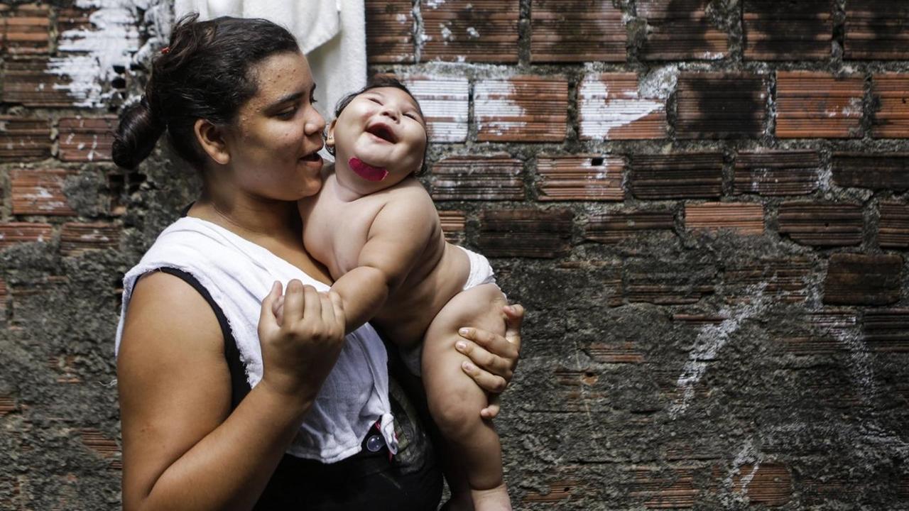 Eine Frau steht vor der Wand eines Hinterhofs und trägt ihr lachendes Kind auf dem Arm