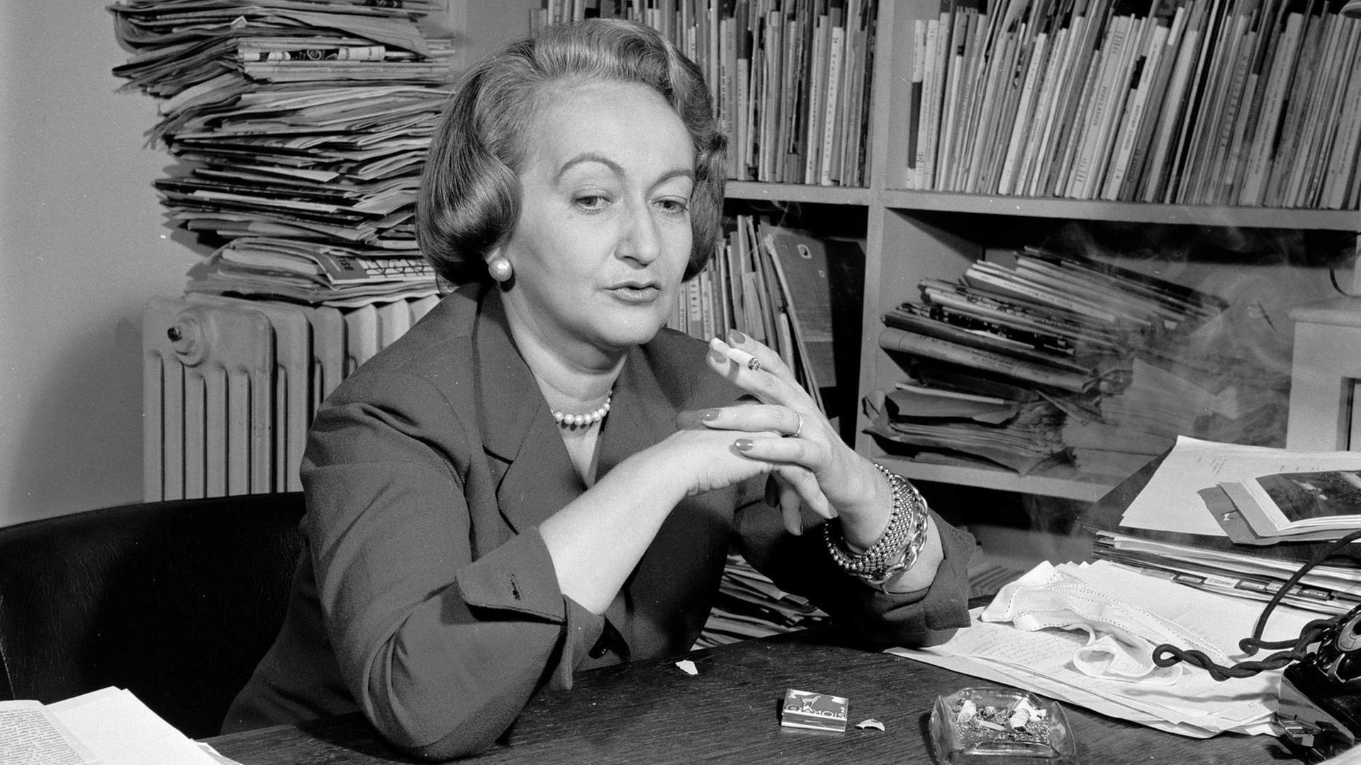 Alba de Céspedes an einem Schreibtisch, mit Zigarette in der Hand vor einem Regal mit vielen Zeitungen