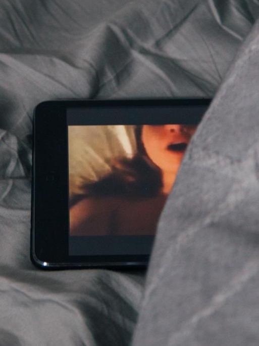 Ein Smartphone-Bildschirm schaut teilweise unter einer Bettdecke hervor. Auf dem Bildschirm ist eine stöhnende Frau beim Sex zu sehen.