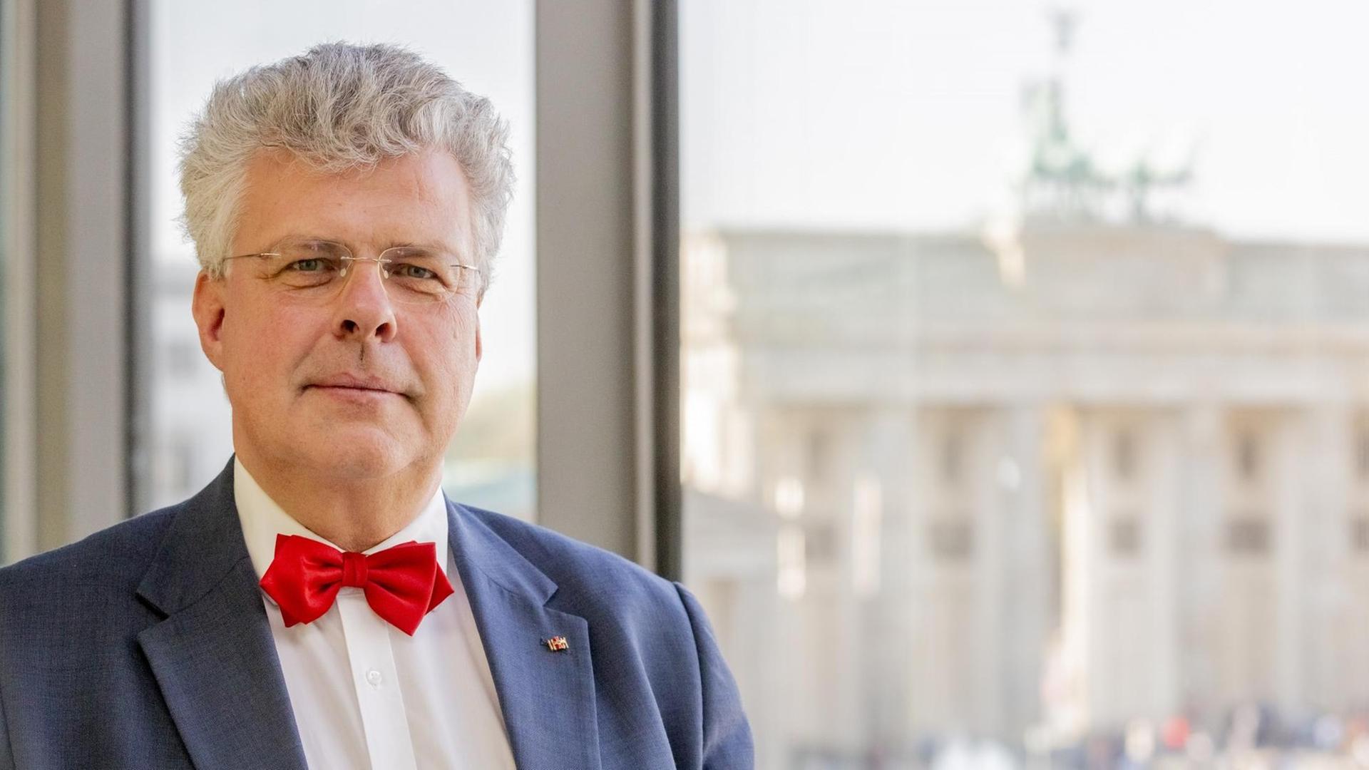 Ein Mann schaut auf einer Porträtaufnahme direkt in die Kamera, er trägt eine rote Fliege, ein weißes Hemd und ein blaues Sakko. Hinter ihm ist eine Fensterfront zu erkennen, durch die man das Brandenburger Tor in Berlin erkennen kann.
