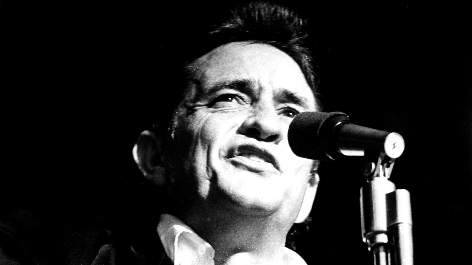 Die Schwarzweiß-Aufnahme zeigt den Sänger Johnny Cash, wie er in ein Mikrofon singt. 
