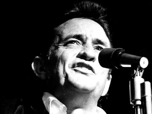 Die Schwarzweiß-Aufnahme zeigt den Sänger Johnny Cash, wie er in ein Mikrofon singt. 
