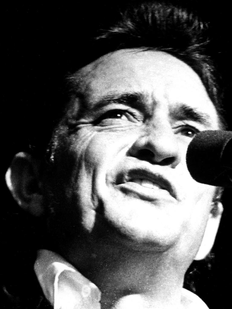 Die Schwarzweiß-Aufnahme zeigt den Sänger Johnny Cash, wie er in ein Mikrofon singt. 