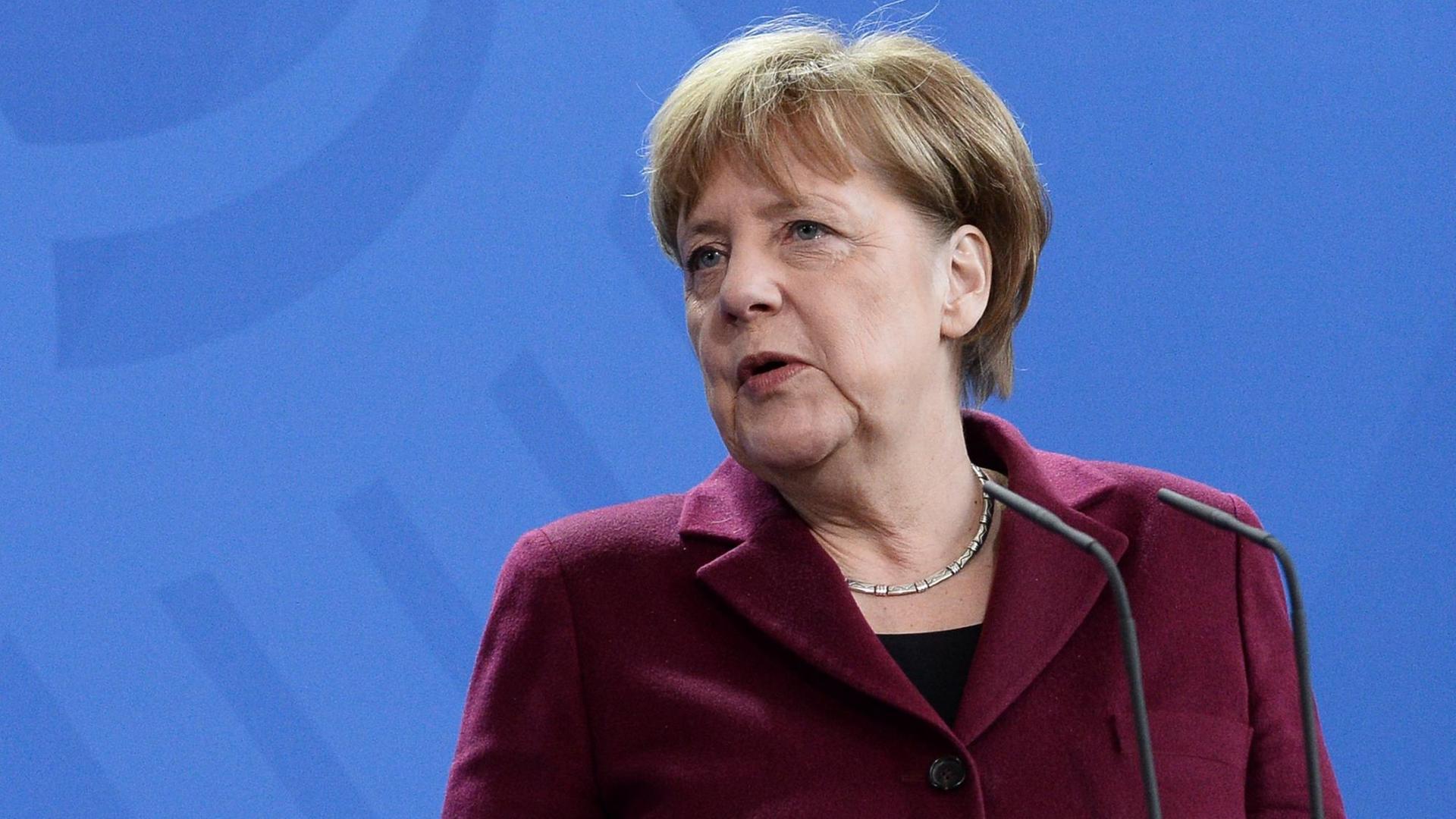 Bundeskanzlerin Angela Merkel in bordeauxrotem Blazer vor blauer Wand mit Bundesadler.
