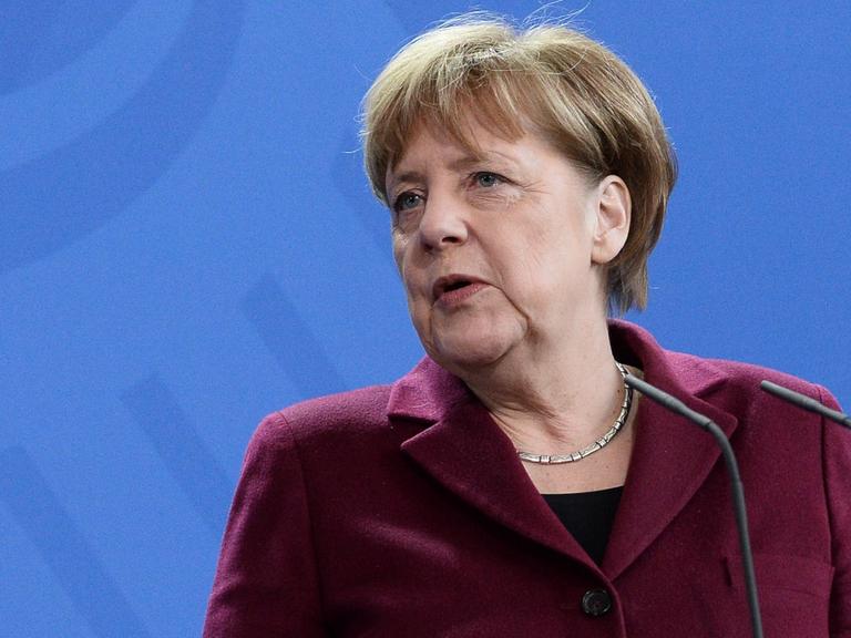 Bundeskanzlerin Angela Merkel in bordeauxrotem Blazer vor blauer Wand mit Bundesadler.