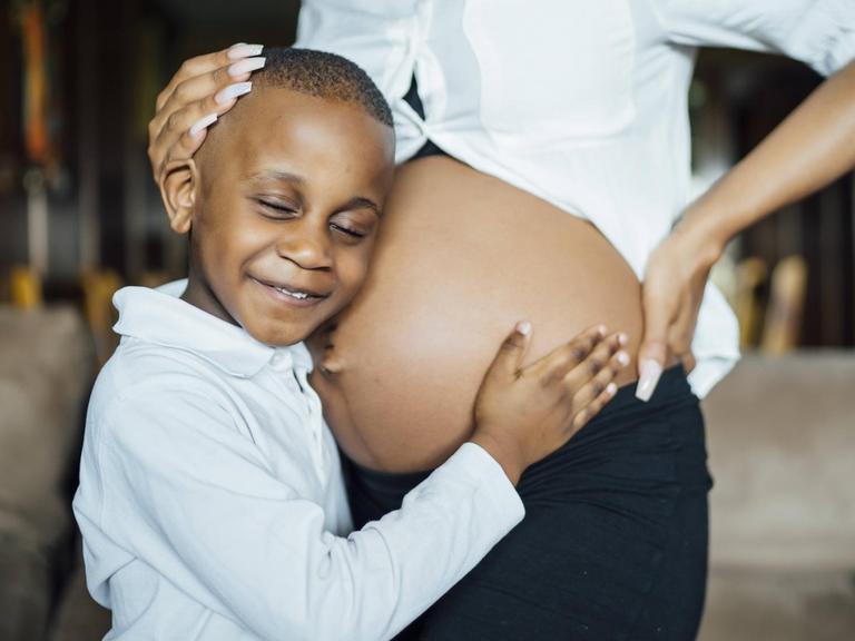 Ein kleiner Junge horcht an dem nackten Bauch einer schwangeren Frau.