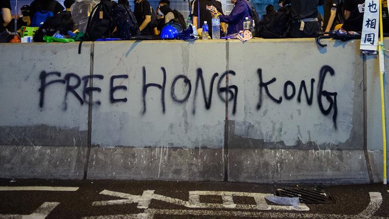 18.08.2019, China, Hongkong: Demonstranten sitzen am Abend auf einer Mauer, die mit einem Grafitti («Free Hong Kong») beschrieben wurde. In der ehemaligen britischen Kronkolonie Hongkong sind die Proteste gegen den Einfluss aus Peking in eine neue Runde gegangen. Die Demokratiebewegung hat zu einer weiteren Großdemonstration aufgerufen. Foto: Gregor Fischer/dpa | Verwendung weltweit