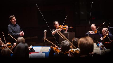 Ein Mann steht vor einem Orchester und singt. Ein Stück neben ihm steht eine Frau, spielt Violine und schaut ebenfalls in Richtung des Orchesters.