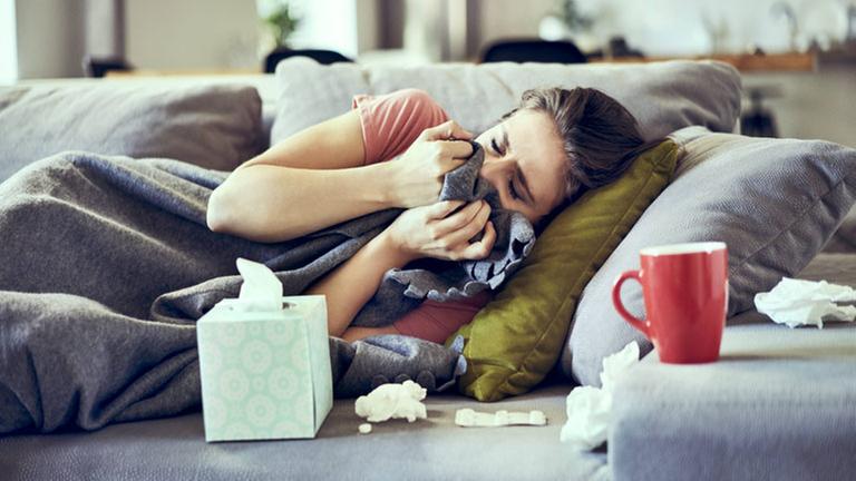 Eine Frau liegt krank auf dem Sofa. Um sie herum Taschentücher und eine Tasse mit Tee. Eine Frau liegt krank auf dem Sofa. Um sie herum Taschentücher und eine Tasse mit Tee.
