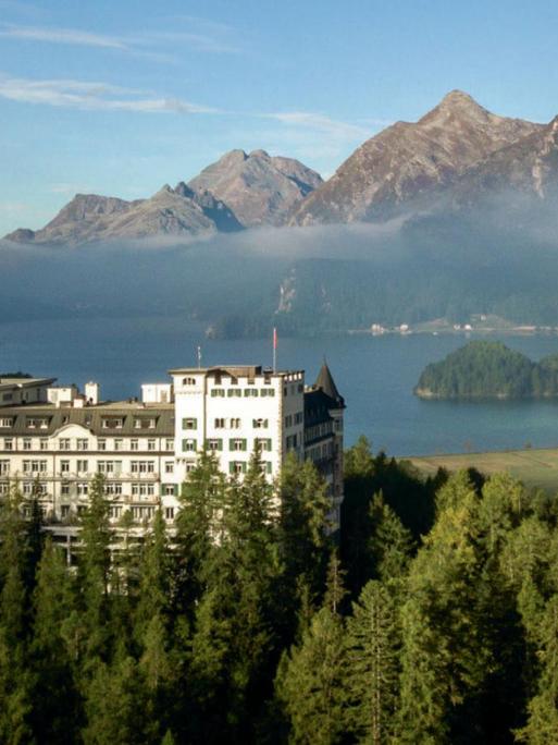 Zu sehen ist das Belle-Époque-Hotel Waldhaus Sils in einer atemberaubenden Umgebung, umrahmt von Bergen, einem See und Wäldern.