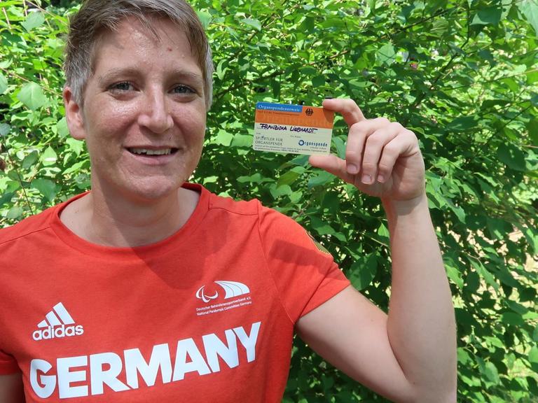 Franziska Liebhardt hält ihren Organspendeausweis in die Höhe. Sie trägt ein rotes T-Shirt mit der Aufschrift Germany.