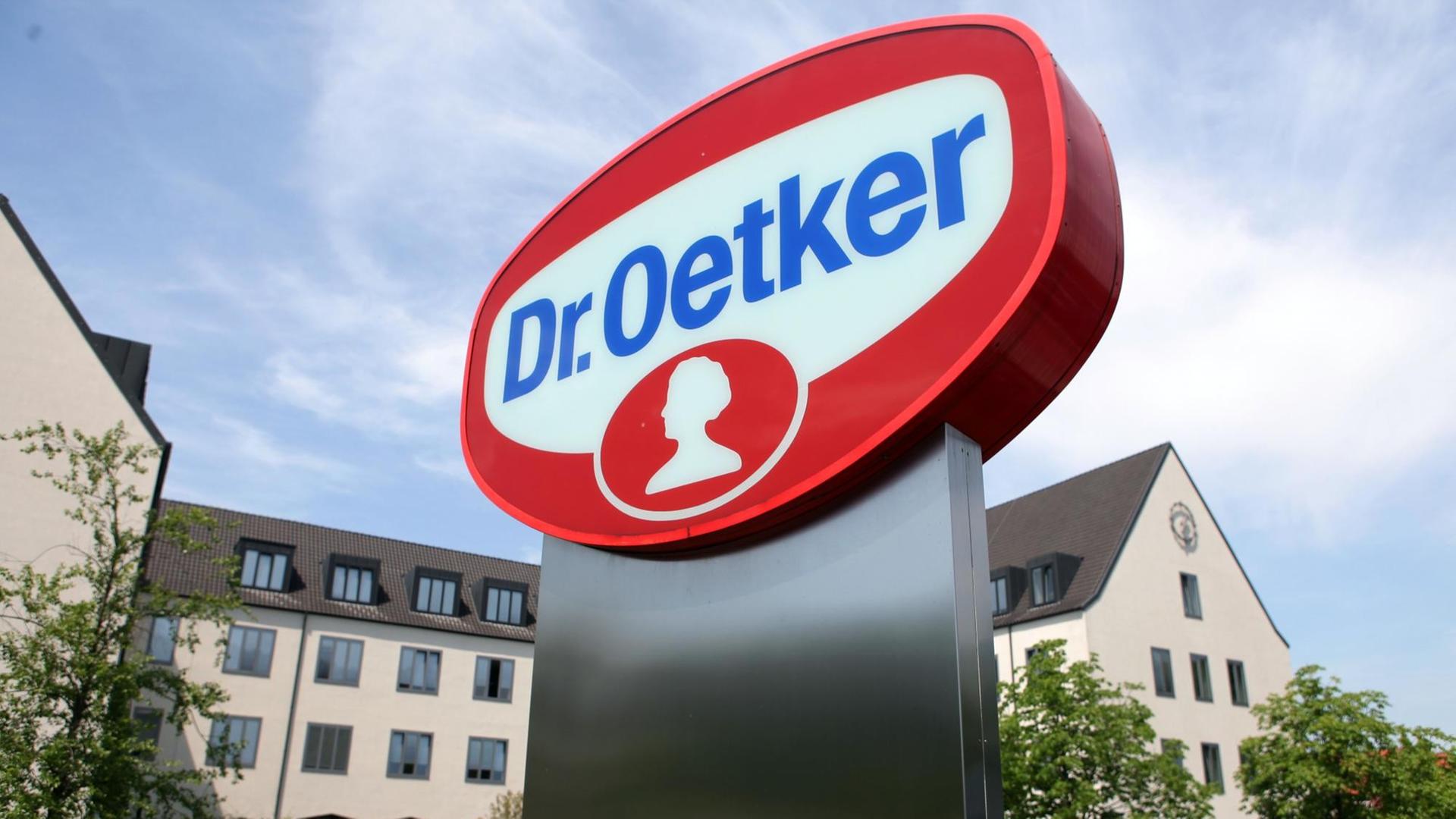 Foto vom Sitz der Dr. August Oetker KG in Bielefeld. Im Vordergrund ist das große Oetker-Logo in den Farben rot, blau und weiß zu sehen.