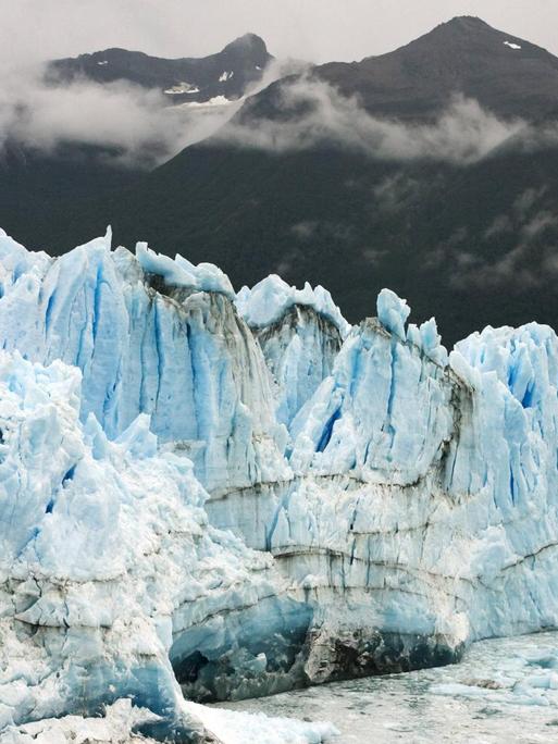 Ein weiß-blau schimmernder Gletscher am Meer im Vordergrund, dunkle Berge im Hintergrund