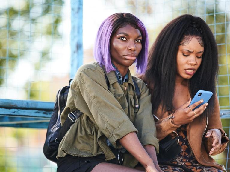 Zwei Frauen sitzen nebeneinander auf einer Bank, eine schaut dabei auf ihr Smartphone, die andere in Richtung der Kamera.
