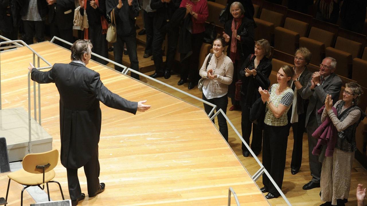 Der Dirigent Mariss Jansons steht ausgebreiteten Armen auf der Bühne, ganz nah an ihm zujubelnden Menschen am Bühnenrand.