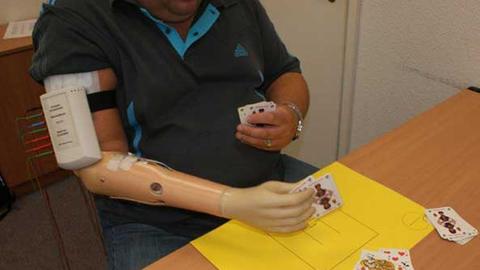Eine Person mit einer Arm-Prothese sitzt an einem Tisch und hält Spielkarten in der Hand.