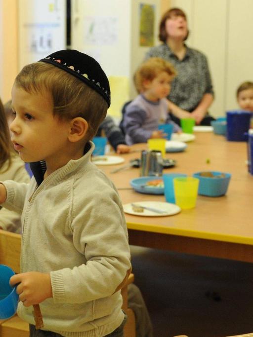 Ein Junge mit einer Kipa trägt einen Teller und einen Becher an einem Tisch vorbei, an dem andere Kinder sitzen.