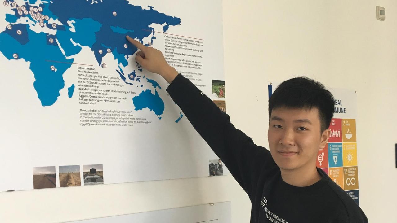 Informatik-Student Eric zeigt stehend auf eine Landkarte, auf der in blau Afrika, Asien und Europa zu sehen ist.