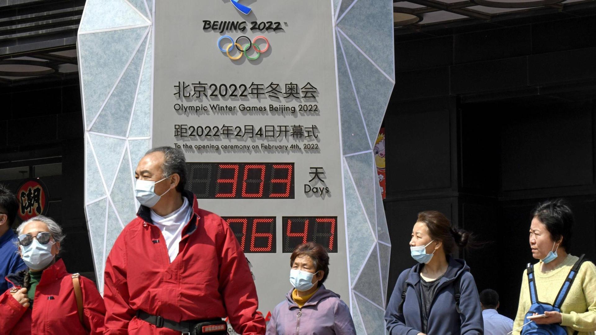 Menschen in Anoraks, mit medizinischen Mund-Nase-Masken zum Schutz vor der Covid19-Pandemie, stehen auf einem Platz in Peking vor einer Uhr mit Digitalanzeige, welche die Zeit bis zum Beginn der olympischen Winterspiele 2022 in Peking herunterzählt.