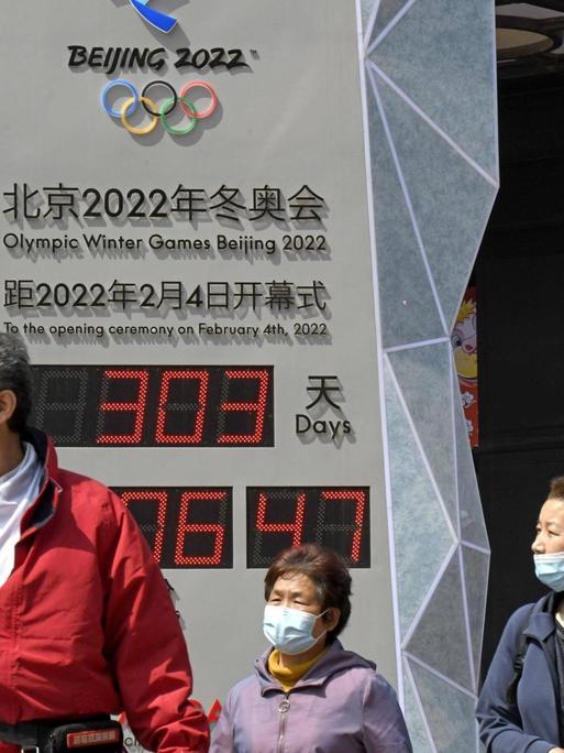 Menschen in Anoraks, mit medizinischen Mund-Nase-Masken zum Schutz vor der Covid19-Pandemie, stehen auf einem Platz in Peking vor einer Uhr mit Digitalanzeige, welche die Zeit bis zum Beginn der olympischen Winterspiele 2022 in Peking herunterzählt.