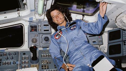 Sally Ride trägt einen hellblauen Raumanzug mit "NASA"-Aufnäher, hinter ihr sind die Schaltknöpfe des Space Shuttle zu sehen.