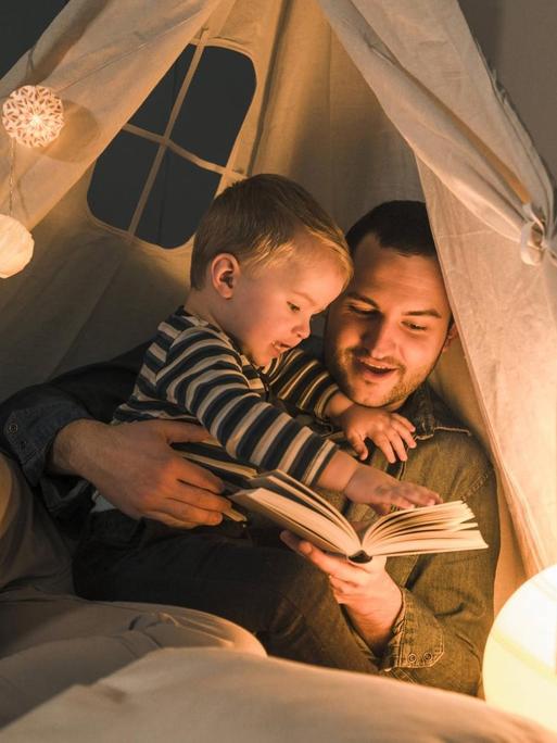 Ein Mann liest seinem Sohn in der Wohnung vor. Beide haben sich in ein Zelt aus weißem Stoff zurückgezogen, das von einer Lampe von innen beleuchtet wird, der Rest der Wohnung ist dunkel.