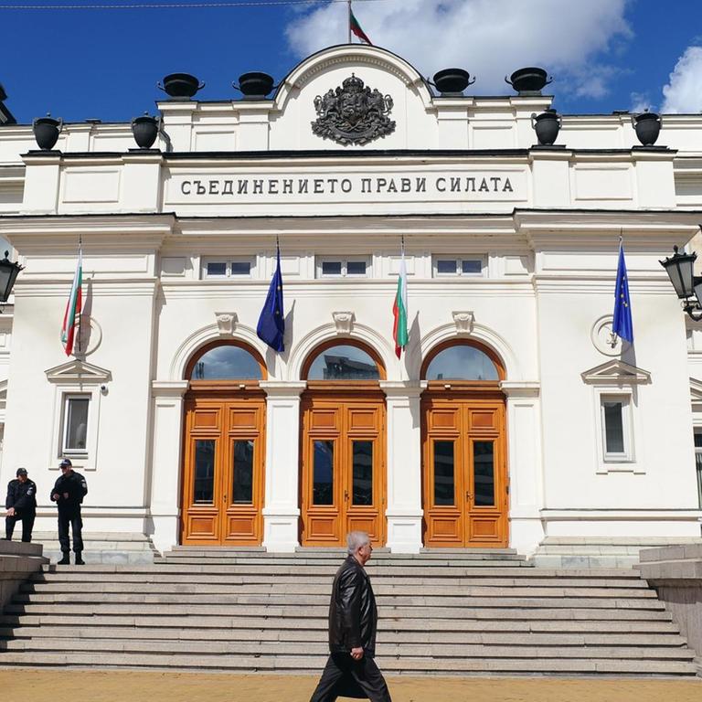 Blick auf die weiße Vorderfront des Parlaments unter wolkig-blauem Himmel. Auf der Treppe zum Eingang zwei Polizisten, ein Mann läuft vor dem Gebäude vorbei.
