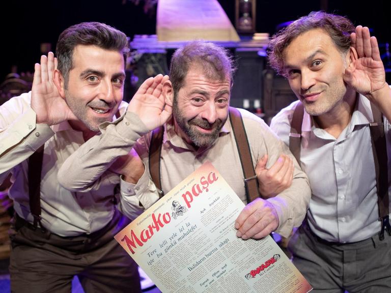 Fatih Koyunoğlu, Erdem Akakçe, Bülent Çolak vom Theater Tiyatroadam gucken in die Kamera und halten jeweils eine Hand hinter die Ohren, als ob sie etwas Bestimmtes hören wollen.