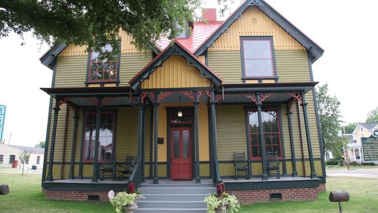 Gelbes Holzhaus mit schwarzen Balken. Das Dach, die Fenster, die Eingangstür und einige Verzierungen sind rot. Vorne befindet sich eine für die Südstaaten typische Veranda.