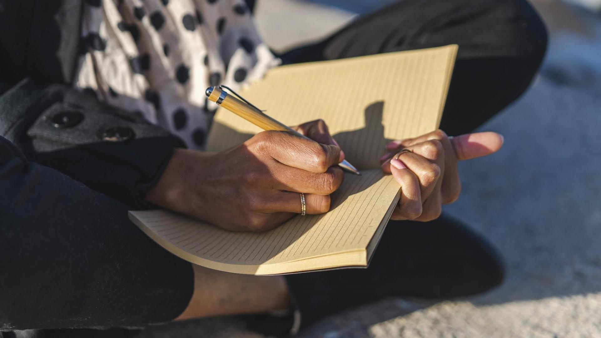 Nahaufnahme von einer Frau, die auf einer Mauer sitzt und in einem Notizbuch schreibt