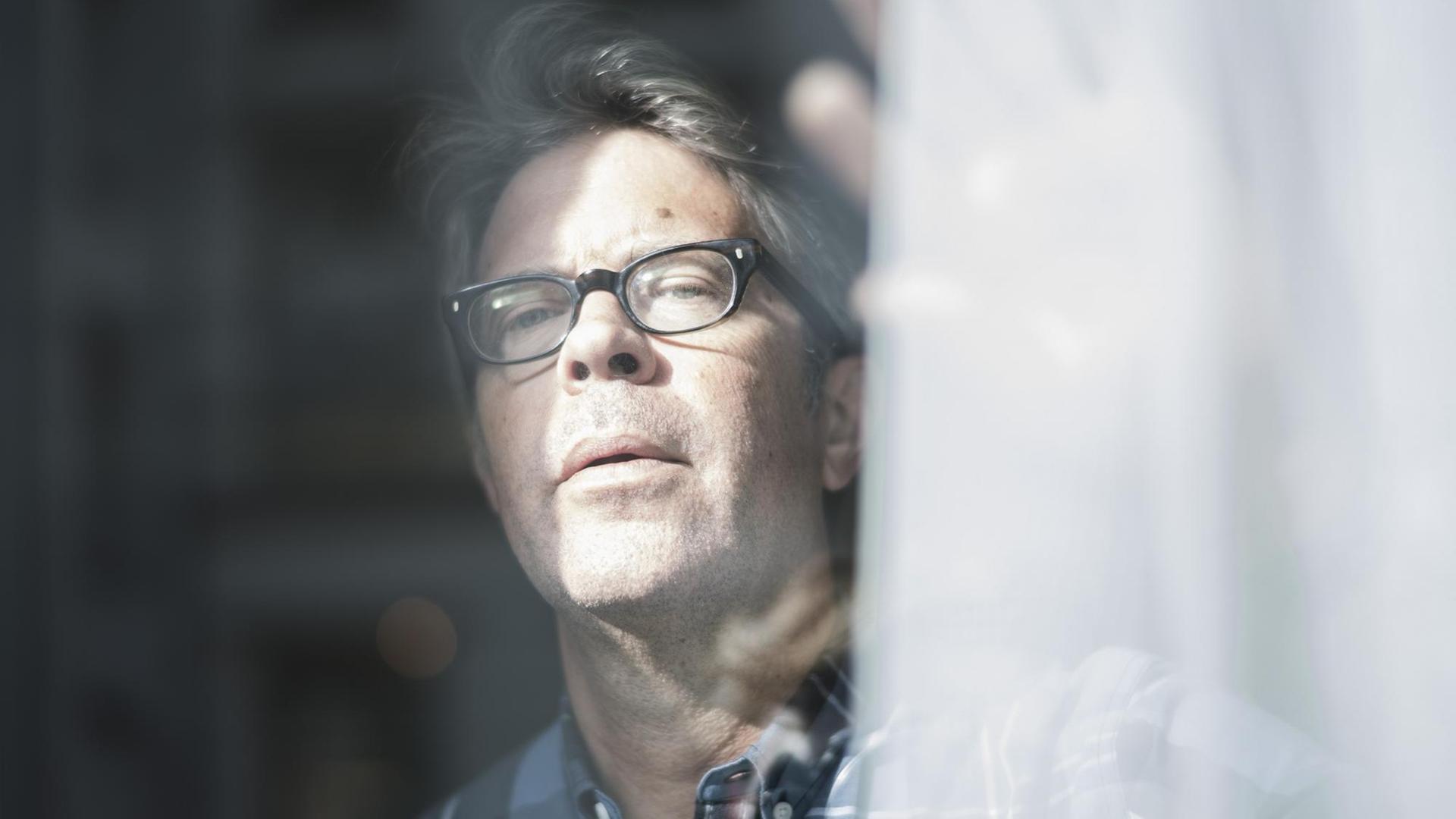 Der US-Autor Jonathan Franzen schaut aus einem Fenster und schiebt dabei eine Gardine beiseite. Er trägt eine Brille.