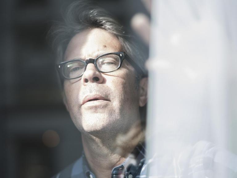 Der US-Autor Jonathan Franzen schaut aus einem Fenster und schiebt dabei eine Gardine beiseite. Er trägt eine Brille.