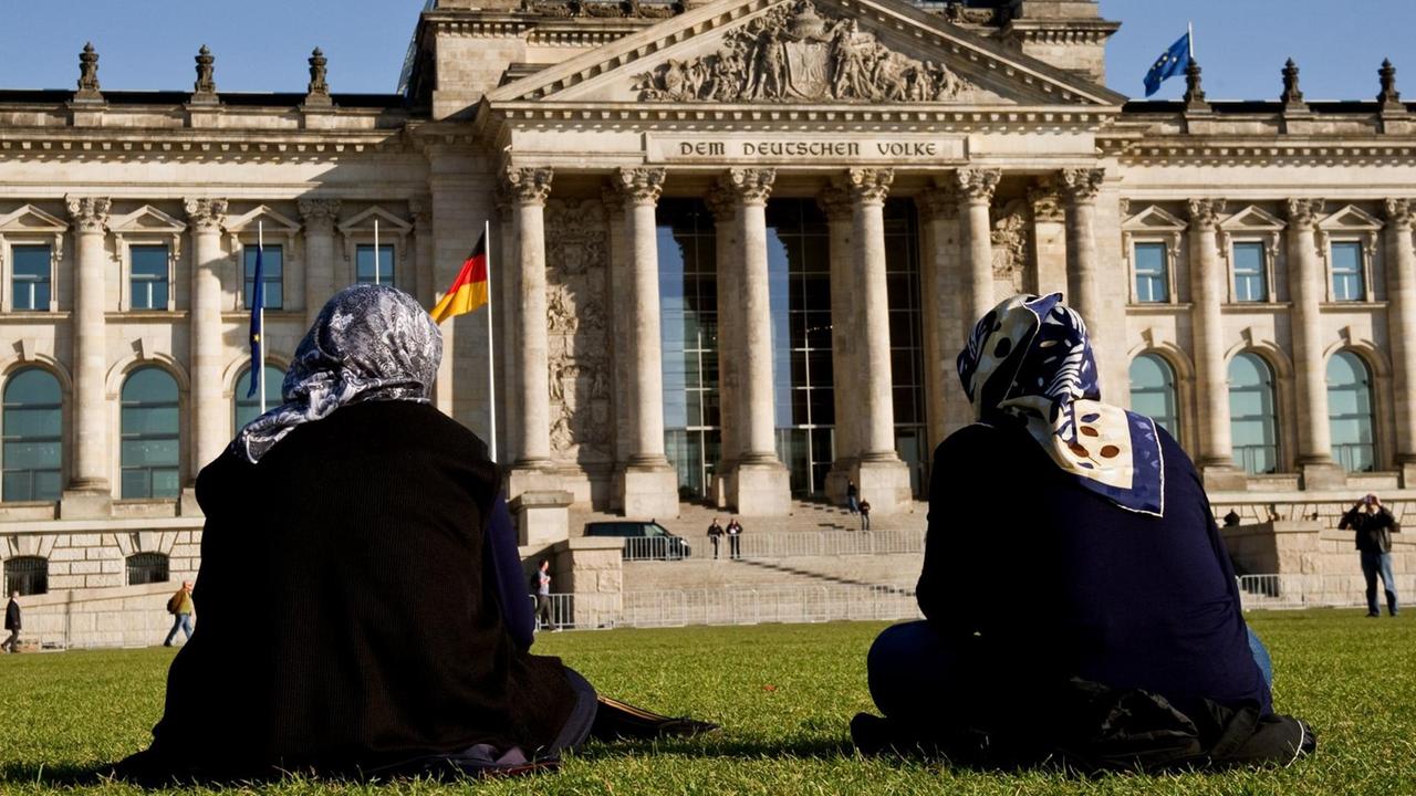 Zwei Frauen mit Kopftuch sitzen auf der Wiese vor dem Reichstagsgebäude in Berlin.