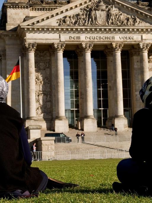 Zwei Frauen mit Kopftuch sitzen auf der Wiese vor dem Reichstagsgebäude in Berlin.