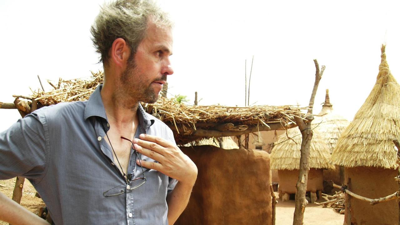 Der Regisseur Christoph Schlingensief steht in Burkina Faso vor mehreren strohgedeckten Lehmhütten.