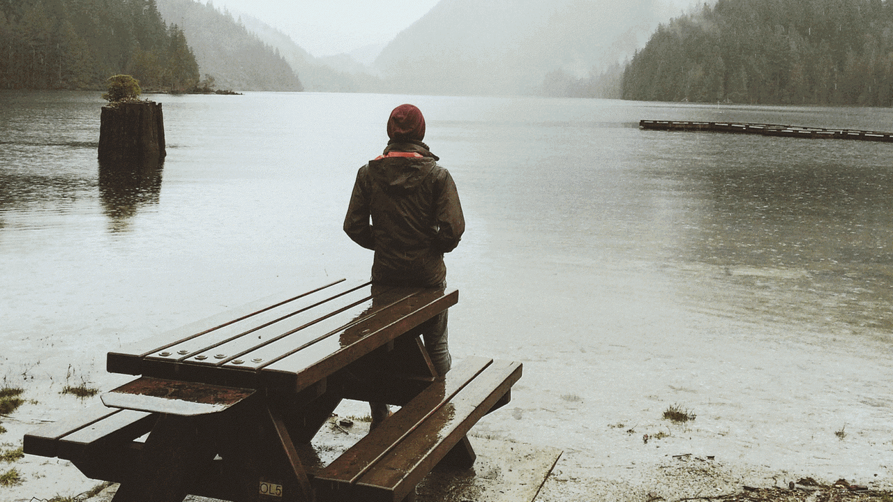 Eine Frau steht alleine vor einem See, mit dem Rücken zur Kamera.