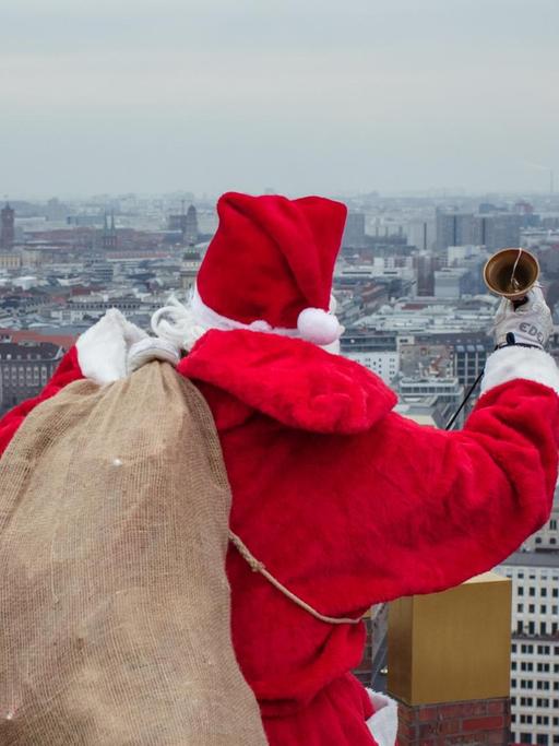 Ein als Weihnachtsmann verkleideter Berufskletterer trägt für eine Werbeaktion auf einem Hochhaus-Dach an Berlins Potsdamer Platz einen Sack voller Geschenke und läutet mit einer Handglocke. Foto: Gregor Fischer/dpa | Verwendung weltweit