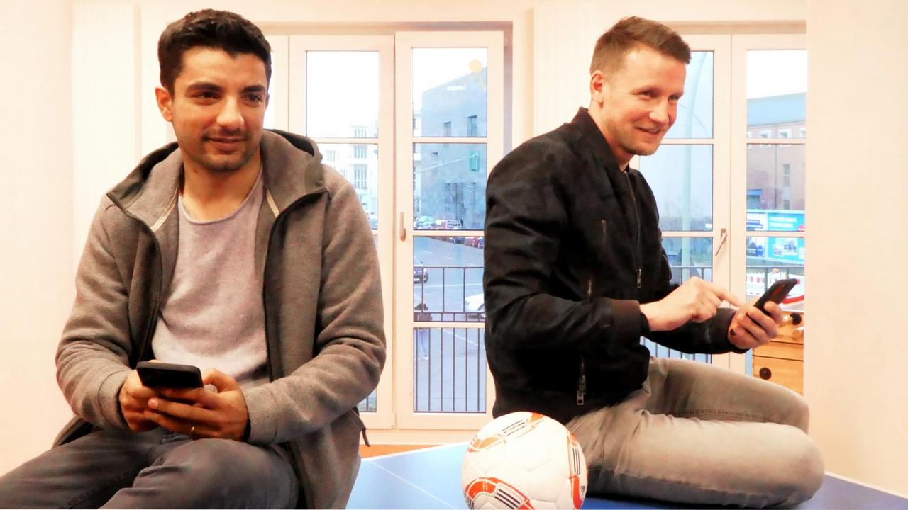 Sami Allagui und Franz Maik vom Hertha BSC sitzen in Alltagskleidung auf einem Tisch. Ein Fußball liegt zwischen ihnen. Sie haben ihre Smartphones in der Hand. 