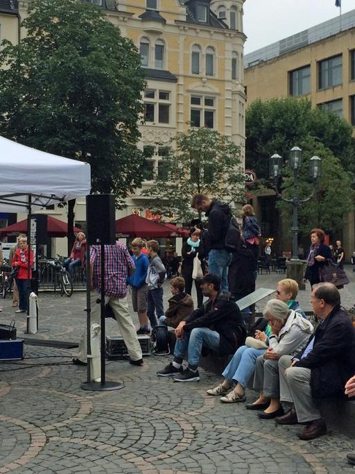 Auf dem Münsterplatz in der Bonner Innenstadt sitzen eine Reihe von Menschen und warten. Ein Flügel ist aufgebaut, überdacht mit einem Zelt gegen den Regen.
