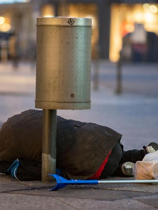 Ein Mann liegt in der Innenstadt unter einem Papierkorb, eine Hand zum Betteln geöffnet.