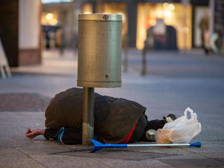 Ein Mann liegt in der Innenstadt unter einem Papierkorb, eine Hand zum Betteln geöffnet.