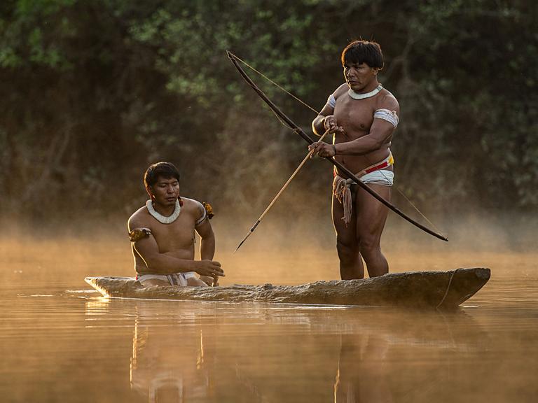 Zwei Männer der indigenen Gruppe Mehinaku auf ihrem flachen Boot auf einem Fluss. Einer der beiden steht und hebt Pfeil und Bogen an. Aus dem Fluss steigt Nebel auf, im Hintergrund scheint die Sonne schwach aufs Wasser.