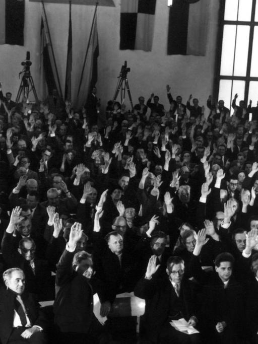 Die Teilnehmer erteilen durch Handzeichen ihre Zustimmung zum Deutschen Manifest. Der historische Saal der Frankfurter Paulskirche war am 29.01.1955 während der Protestkundgebung mit den Flaggen aller deutschen Länder geschmückt. Auf Initiative der SPD und des Deutschen Gewerkschaftsbundes (DGB) hatten sich Persönlichkeiten des öffentlichen Lebens eingefunden, um gegen die Ratifizierung der Pariser Verträge zu protestieren. Zum Schluss der Kundgebung nahmen sie einstimmig ein Manifest ("Deutsches Manifest") an, in dem Volk und Regierung zum Widerstand gegen die sich immer stärker werdende Tendenz zur Spaltung Deutschland aufgerufen wurden.
