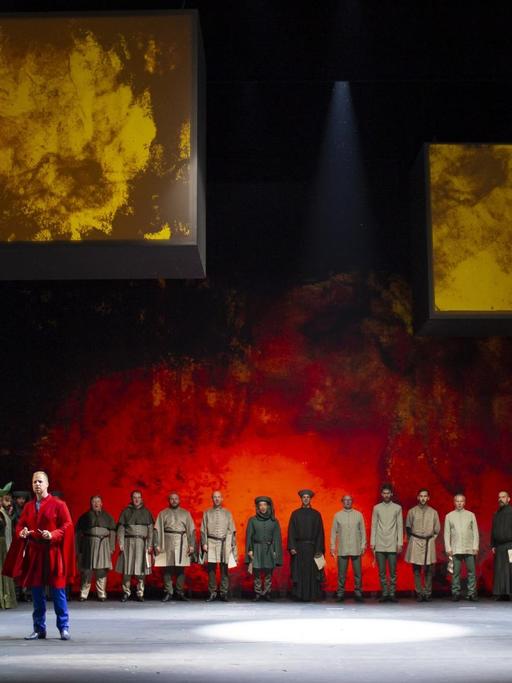 Auf einer rot erleuchteten Bühne steht ein Chor in historischer Kleidung im Halbrund um einen einzelnen Protagonisten.