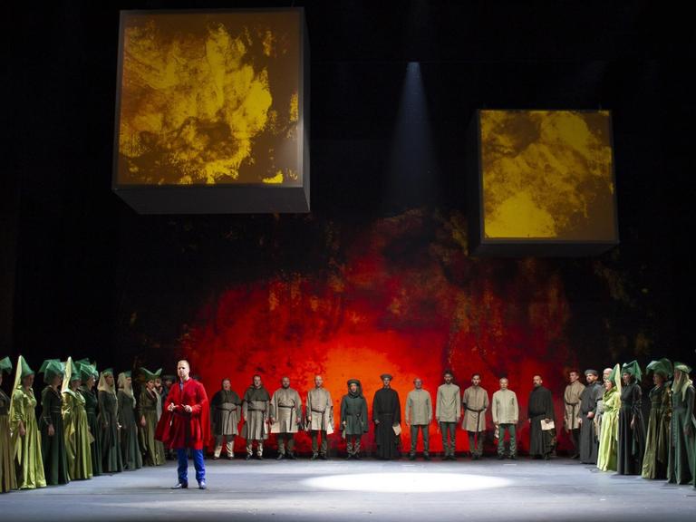 Auf einer rot erleuchteten Bühne steht ein Chor in historischer Kleidung im Halbrund um einen einzelnen Protagonisten.