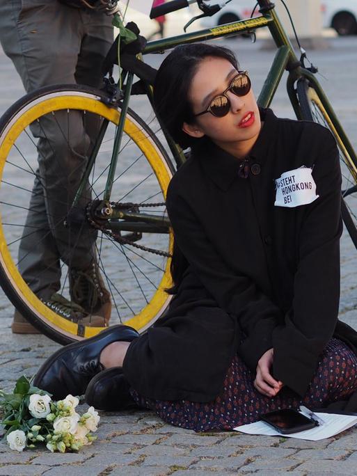 Die Künstlerin Alice Kahei Yu aus Hongkong sitzt hinter einem Blumenstrauß auf dem Pflaster, sie trägt eine Sonnenbrille und auf ihrem Pullover steht "Steht Hongkong bei"
