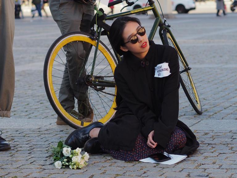 Die Künstlerin Alice Kahei Yu aus Hongkong sitzt hinter einem Blumenstrauß auf dem Pflaster, sie trägt eine Sonnenbrille und auf ihrem Pullover steht "Steht Hongkong bei"